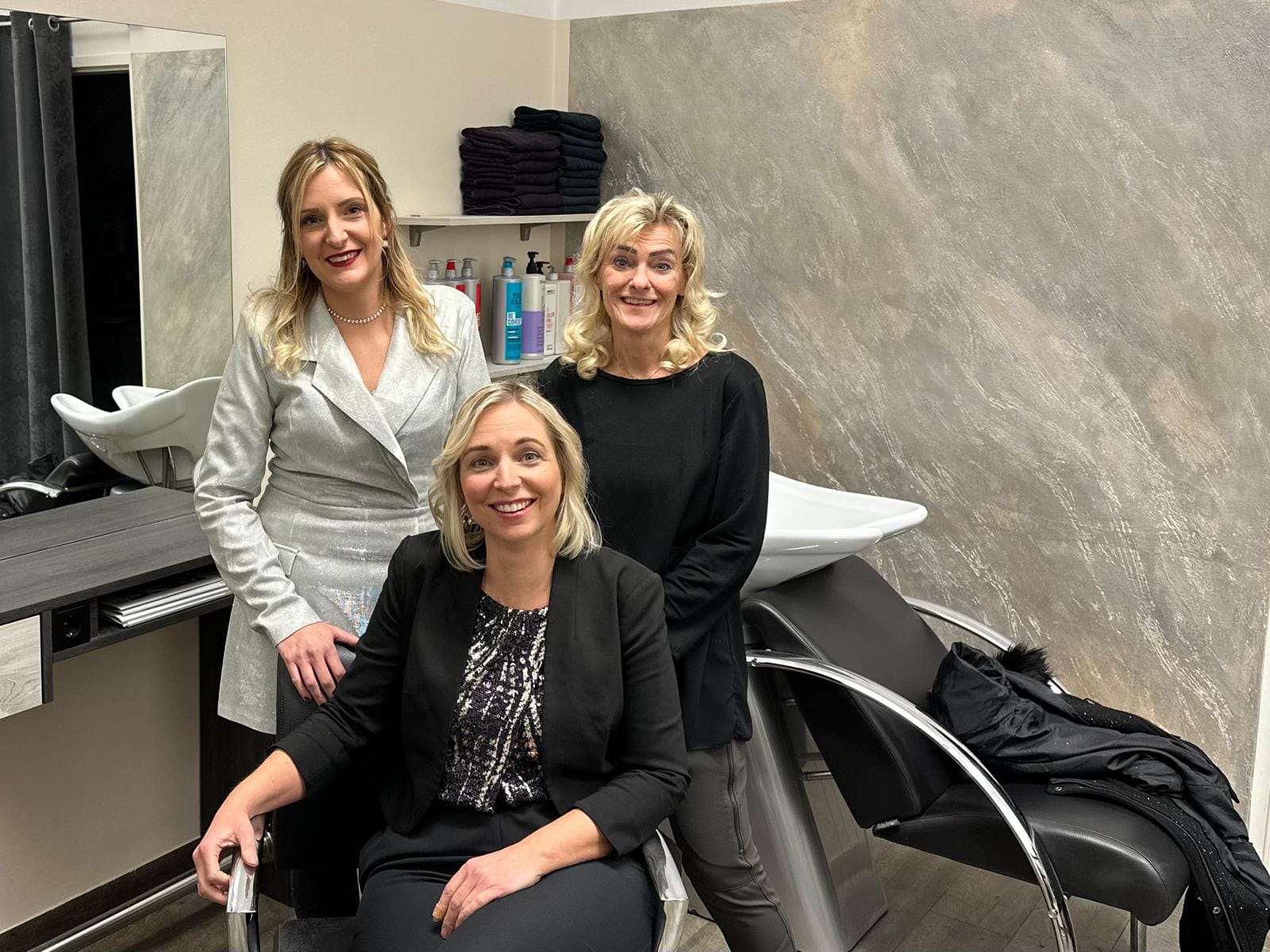 Ein Bild von drei Friseuren im Haarstudio Style, Klagenfurt. Die Inhaberin Frau Cornelia Buchsbaum ist in der Mitte, flankiert von zwei Kolleginnen. Das Studio ist modern eingerichtet mit einem großen Spiegel, Regalen voller Pflegeprodukte und stylischen Stühlen. Das Haarstudio Style ist bekannt für seine hochwertigen Dienstleistungen wie Haarverlängerungen und Extensions in Klagenfurt. Ein Ort, an dem Mode und professionelle Haarbehandlung auf höchstem Niveau aufeinandertreffen.